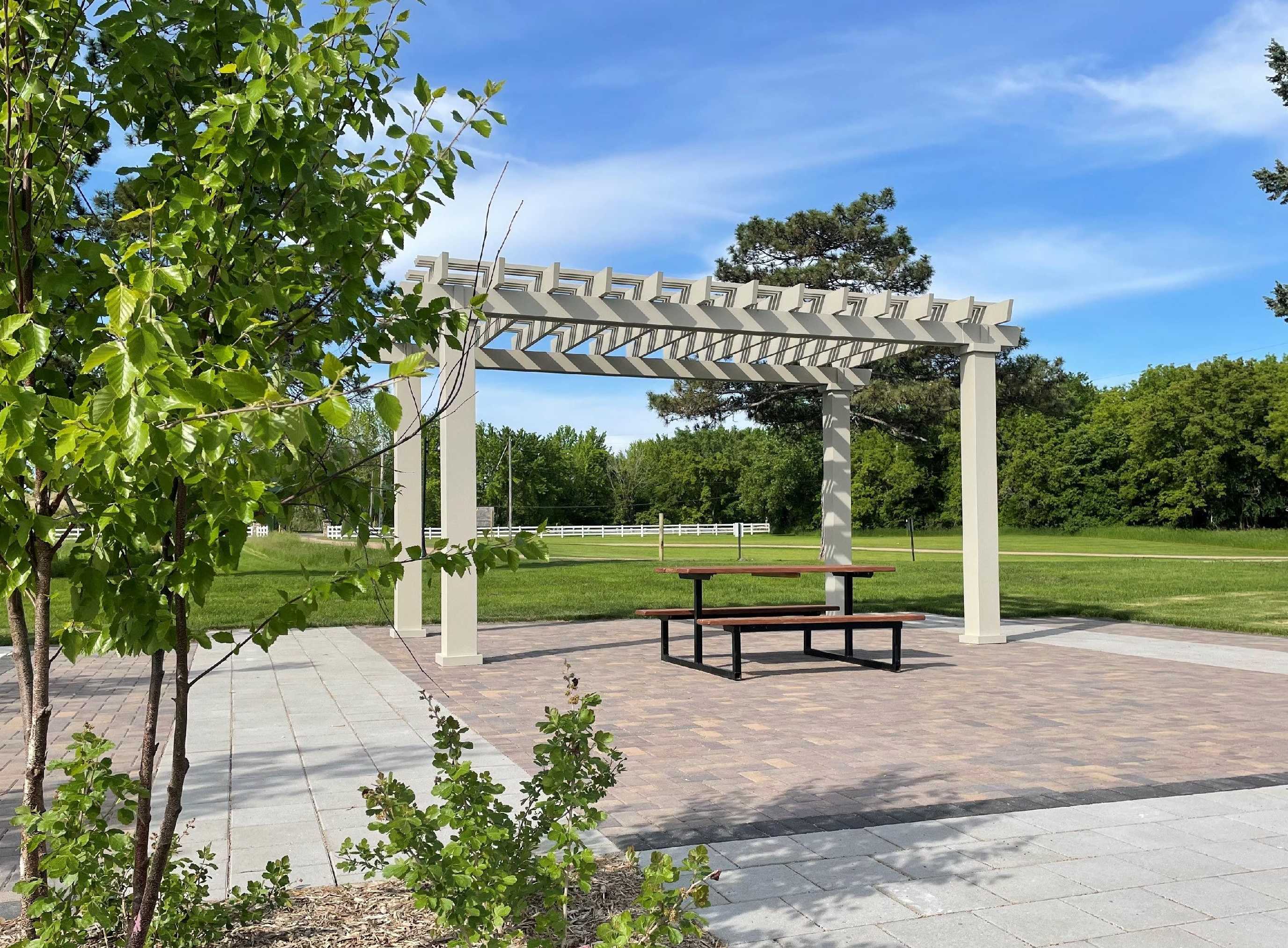 A serene park scene with a white pergola over a picnic table on a paved area. Lush green trees and grass stretch into the distance under a blue sky. - Copy